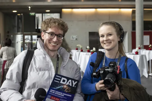 2024: Zwei Teilnehmende mit Kamera und eDay-Flyer im Foyer beim eDay Salzburg.