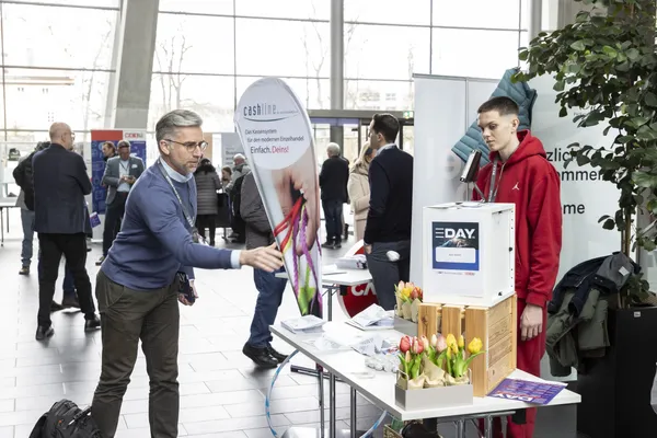 2024: Besucher interagiert mit einem Ausstellerstand mit Infomaterial und Blumen.