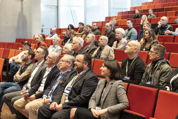 2025: Publikum sitzt in roten Sitzen und hört Vorträgen im Saal.