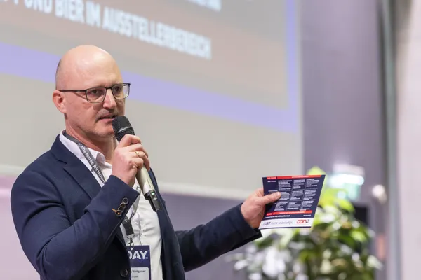 2025: Vortragende Person hält einen Flyer und spricht auf der Bühne.