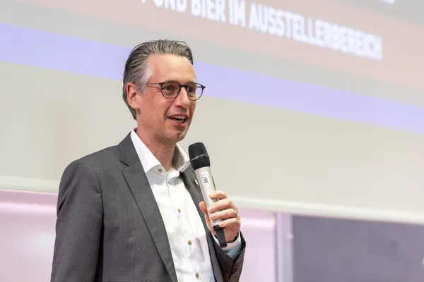 2025: Vortragende Person spricht auf der Bühne vor projizierter Präsentation.