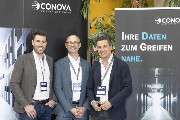 Drei Personen vor CONOVA-Messestand mit Bannern und Badges beim eDay Salzburg 2025