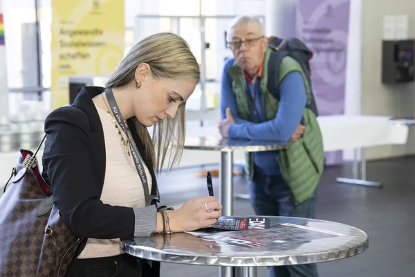 Teilnehmerin trägt sich an Stehtisch ein, im Hintergrund Personen und Messestände beim eDay Salzburg 2025