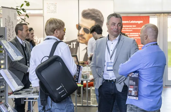 Teilnehmende unterhalten sich vor Messestand mit großen Bannerbildern beim eDay Salzburg 2025