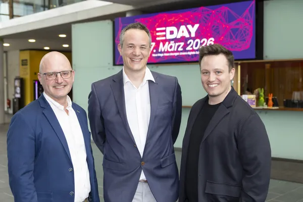 Drei Personen posieren vor E‑DAY-Banner 'März 2026' in der Empfangshalle beim eDay Salzburg 2026.